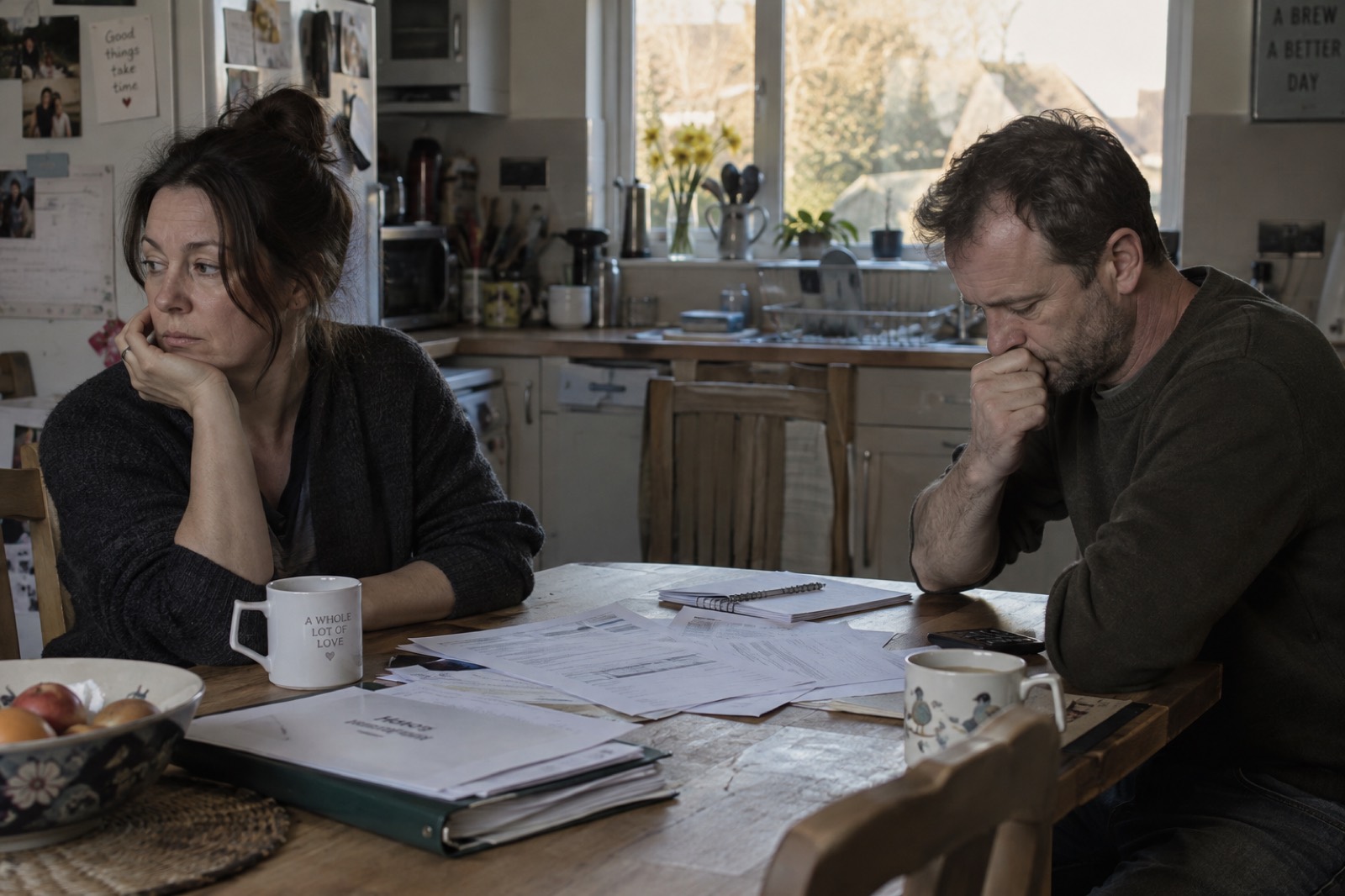 A couple at their kitchen table with paperwork spread between them, both looking away, neither speaking.