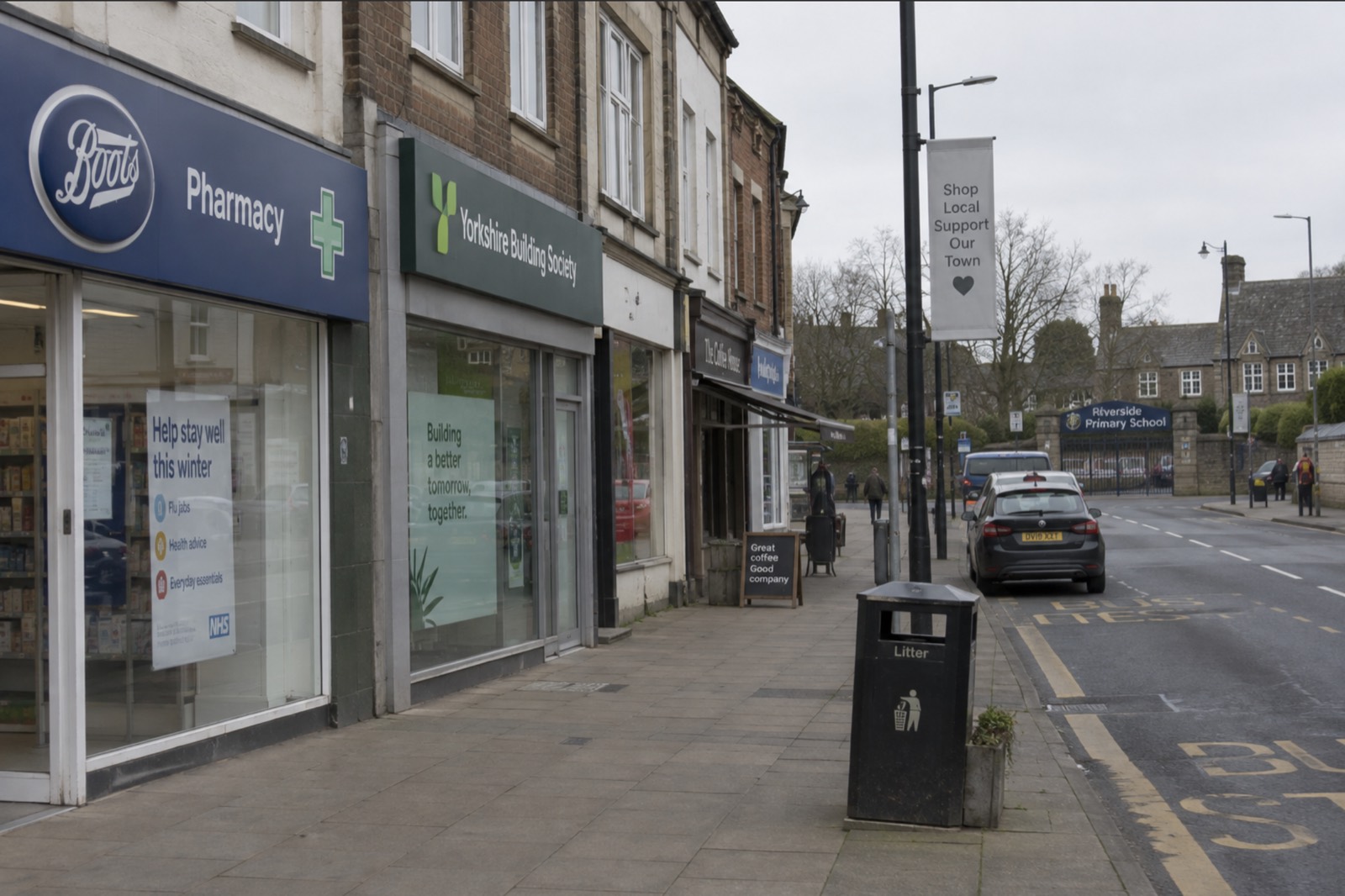An ordinary British high street on a weekday morning: a pharmacy, a building society, a coffee shop, and a primary school further along the road.